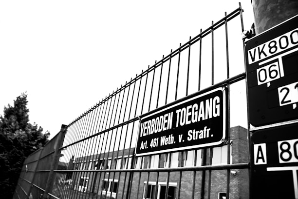 A black and white image of a metal fence with a sign reading 'Verboden Toegang' in Dutch, translating to 'No Trespassing'. The sign references a specific legal article. In the background, part of a building and a tree are visible.