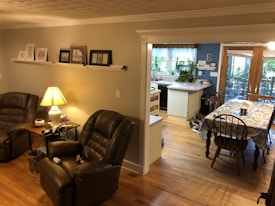 A cozy living space with two brown leather recliners positioned around a wooden side table with a lamp. The walls are decorated with framed pictures on a long shelf. The room transitions into a dining area with a floral tablecloth on a wooden table, set near large windows that bring in natural light. In the background, there's a kitchen area with white cabinets, plants, and a window overlooking greenery.