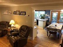 A cozy living space with two brown leather recliners positioned around a wooden side table with a lamp. The walls are decorated with framed pictures on a long shelf. The room transitions into a dining area with a floral tablecloth on a wooden table, set near large windows that bring in natural light. In the background, there's a kitchen area with white cabinets, plants, and a window overlooking greenery.