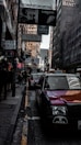 A bustling urban street with tall buildings adorned with numerous signs and advertisements. Several taxis are lined up on the road, and pedestrians are walking along the sidewalk. The architecture is dense, with various shops and hotels visible in the background.