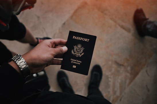 A person is holding a United States passport in their hand. The hand is wearing a silver watch, and the person is standing on a tile floor. There are a pair of shoes visible, suggesting the person is looking down. The image has a warm tone with subtle lighting.