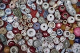 A collection of various ceramic and porcelain pots and vases are arranged closely together on a red surface. The items vary in color and size, including patterns of blue and white, floral designs, and earthy tones. Some objects have intricate decorations, while others are plain. There are also unique shapes, such as animal figurines and masks.