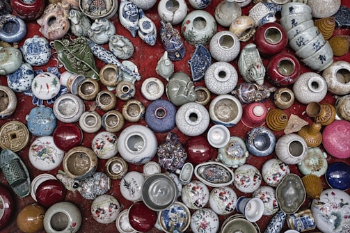 A collection of various ceramic and porcelain pots and vases are arranged closely together on a red surface. The items vary in color and size, including patterns of blue and white, floral designs, and earthy tones. Some objects have intricate decorations, while others are plain. There are also unique shapes, such as animal figurines and masks.