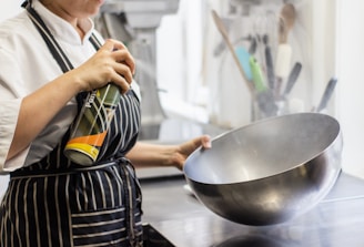 A person wearing a white chef's uniform is using a can of spray on a large metallic bowl. The bowl is being held with one hand, and a spray can with a partial label in green, yellow, and orange is in the other hand. The background shows kitchen utensils, including spatulas and ladles, in various colors.