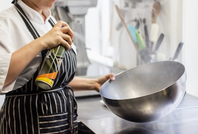 A person wearing a white chef's uniform is using a can of spray on a large metallic bowl. The bowl is being held with one hand, and a spray can with a partial label in green, yellow, and orange is in the other hand. The background shows kitchen utensils, including spatulas and ladles, in various colors.