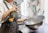 A person wearing a white chef's uniform is using a can of spray on a large metallic bowl. The bowl is being held with one hand, and a spray can with a partial label in green, yellow, and orange is in the other hand. The background shows kitchen utensils, including spatulas and ladles, in various colors.