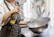 A person wearing a white chef's uniform is using a can of spray on a large metallic bowl. The bowl is being held with one hand, and a spray can with a partial label in green, yellow, and orange is in the other hand. The background shows kitchen utensils, including spatulas and ladles, in various colors.