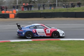 A race car with a blue and red color scheme speeds along a track, bearing the number 8 and various sponsor logos, including Valluga. In the background, spectators in orange vests stand behind a barrier watching the event.
