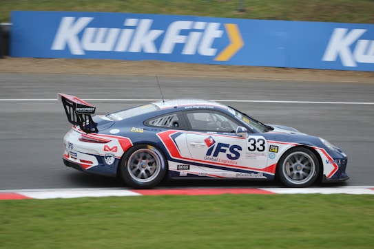 A race car with the number 33 and sporting FS Global Logistics branding speeds along a racetrack. The car is painted in blue, white, and red. The background shows a Kwik Fit advertisement banner next to a grassy area.