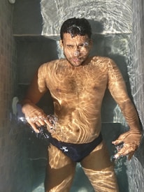 A person is floating horizontally in a swimming pool with their face above water. Sunlight creates dynamic patterns on the water's surface and the person's skin. The person appears relaxed, with some air bubbles around their face.