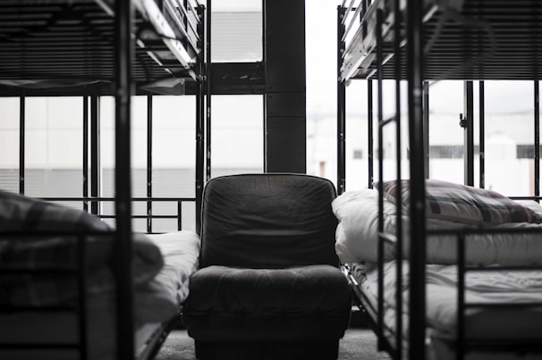 A dormitory setting with unoccupied bunk beds lined up along the walls, surrounding a central armchair. The room appears neat and orderly, with folded blankets on the beds. Large windows allow natural light to illuminate the space, adding a slightly somber and quiet atmosphere.
