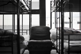 A dormitory setting with unoccupied bunk beds lined up along the walls, surrounding a central armchair. The room appears neat and orderly, with folded blankets on the beds. Large windows allow natural light to illuminate the space, adding a slightly somber and quiet atmosphere.