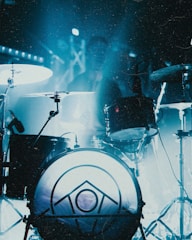 A drum set is illuminated by blue lighting, creating an atmospheric scene. The foreground features a drum with a geometric pattern on its surface, while cymbals and other drum components are visible. A figure is somewhat obscured in the background, likely a drummer.