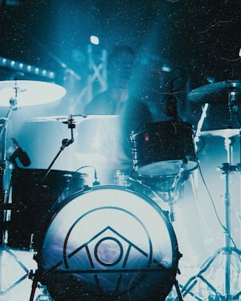 A drum set is illuminated by blue lighting, creating an atmospheric scene. The foreground features a drum with a geometric pattern on its surface, while cymbals and other drum components are visible. A figure is somewhat obscured in the background, likely a drummer.
