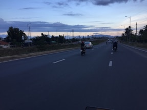 A long, open road with a few motorcycles and cars traveling in the same direction. The sky has a subtle sunset with clouds scattered across the horizon. The sides of the road are lined with trees and some buildings. Streetlights line the highway, and there are mountains visible in the distance.