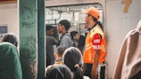 A group of people is boarding a crowded train, with some standing inside. A person wearing an orange high-visibility jacket and cap, possibly a train official, is monitoring the situation near the door. Passengers are seen carrying bags and appear to be in transit.