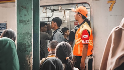 A group of people is boarding a crowded train, with some standing inside. A person wearing an orange high-visibility jacket and cap, possibly a train official, is monitoring the situation near the door. Passengers are seen carrying bags and appear to be in transit.