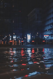 A nighttime city scene with tall buildings and bright lights reflecting off a water surface. The lights are predominantly red and blue, creating a vibrant and slightly mysterious atmosphere. The scene is likely urban, with concrete structures lining either side of the image.