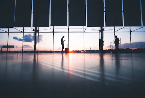 Silhouettes of people with luggage in a terminal against a bright low sun