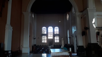 A large, dimly-lit church interior with arched windows creating intricate patterns of light. A statue is positioned on the right side of the altar, which is covered with a decorative cloth. Tall columns and high ceilings create a spacious and majestic atmosphere.