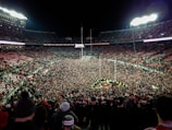 A massive crowd fills a sports stadium after an event, with people covering the field and the stands partially occupied. Bright stadium lights illuminate the scene, creating a vibrant atmosphere as individuals celebrate together.