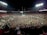A massive crowd fills a sports stadium after an event, with people covering the field and the stands partially occupied. Bright stadium lights illuminate the scene, creating a vibrant atmosphere as individuals celebrate together.
