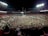 A massive crowd fills a sports stadium after an event, with people covering the field and the stands partially occupied. Bright stadium lights illuminate the scene, creating a vibrant atmosphere as individuals celebrate together.