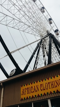 A large Ferris wheel with white and black structure is prominently visible against a cloudy sky. Below it, a sign reads 'African Clothing' with a decorative border, indicating a shop or store.