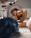 A figurine of a baseball player in a uniform is next to a trophy shaped like a baseball glove holding a baseball. The background displays a blurred sports team logo, and in the foreground are a blurred baseball cap and a baseball.