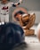 A figurine of a baseball player in a uniform is next to a trophy shaped like a baseball glove holding a baseball. The background displays a blurred sports team logo, and in the foreground are a blurred baseball cap and a baseball.