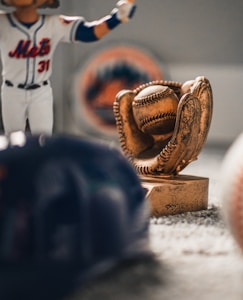 A figurine of a baseball player in a uniform is next to a trophy shaped like a baseball glove holding a baseball. The background displays a blurred sports team logo, and in the foreground are a blurred baseball cap and a baseball.