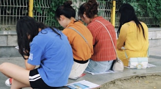 Four diverse girls sitting together under a tree, sharing a book and laughing.