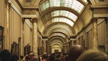 A large gallery hall features ornate arches and decorative columns with intricate designs. The space is filled with numerous people, indicating a busy and popular setting. Framed artworks line the walls of the hallway, which extends far into the distance.