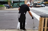A person wearing black clothing and gloves is cleaning the street using a litter picker tool. They are focusing on a drainage area near a sidewalk. In the background, there is a busy street with cars, a bus, and a store with large windows.