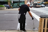 A person wearing black clothing and gloves is cleaning the street using a litter picker tool. They are focusing on a drainage area near a sidewalk. In the background, there is a busy street with cars, a bus, and a store with large windows.