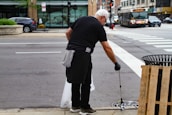A person wearing black clothing and gloves is cleaning the street using a litter picker tool. They are focusing on a drainage area near a sidewalk. In the background, there is a busy street with cars, a bus, and a store with large windows.