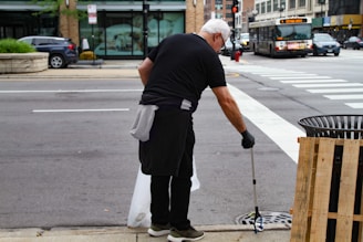 A person wearing black clothing and gloves is cleaning the street using a litter picker tool. They are focusing on a drainage area near a sidewalk. In the background, there is a busy street with cars, a bus, and a store with large windows.