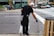 A person wearing black clothing and gloves is cleaning the street using a litter picker tool. They are focusing on a drainage area near a sidewalk. In the background, there is a busy street with cars, a bus, and a store with large windows.