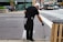 A person wearing black clothing and gloves is cleaning the street using a litter picker tool. They are focusing on a drainage area near a sidewalk. In the background, there is a busy street with cars, a bus, and a store with large windows.