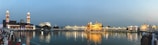 A twilight scene featuring a beautifully illuminated golden building with its reflection shimmering in a calm water body. Tall towers with intricate designs stand nearby, and the sky transitions from a soft blue to a gentle orange. People gather along the water's edge, some wearing traditional attire, enhancing the cultural atmosphere.