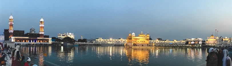 A twilight scene featuring a beautifully illuminated golden building with its reflection shimmering in a calm water body. Tall towers with intricate designs stand nearby, and the sky transitions from a soft blue to a gentle orange. People gather along the water's edge, some wearing traditional attire, enhancing the cultural atmosphere.