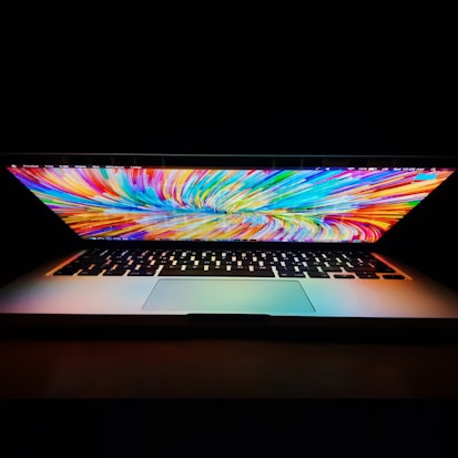 A partially open laptop displays a vivid, colorful abstract pattern on the screen. The backdrop is dark, highlighting the laptop's illuminated keyboard and vibrant screen.