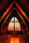 The interior of a church with a high, arched wooden ceiling and rows of pews leading to an altar. The focal point is a large, backlit stained glass window featuring a cross with a crucifix. The warm lighting gives the scene a serene and contemplative atmosphere.