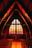 The interior of a church with a high, arched wooden ceiling and rows of pews leading to an altar. The focal point is a large, backlit stained glass window featuring a cross with a crucifix. The warm lighting gives the scene a serene and contemplative atmosphere.