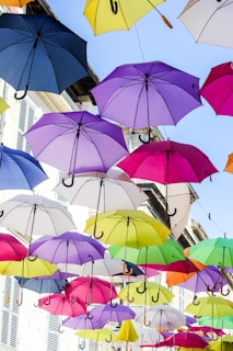 Multiple colorful umbrellas are suspended in the air above a street, creating a canopy effect. The umbrellas come in various colors including blue, purple, pink, yellow, and green. Behind the umbrellas, there are the facades of buildings with white shutters on the windows.