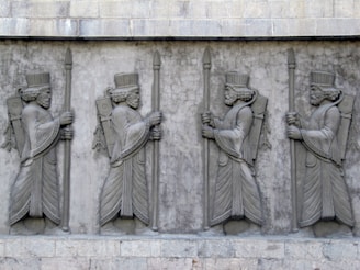 A stone relief sculpture depicting four identical figures in ancient attire, each holding a staff. They are adorned with elaborate headdresses and robes, featuring intricate patterns and details typical of an ancient civilization. The figures are lined up in a row, displaying symmetry and exuding a sense of regality.