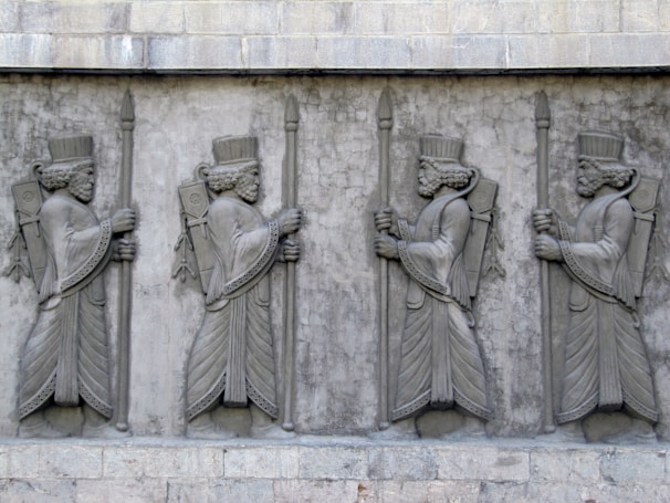 A stone relief sculpture depicting four identical figures in ancient attire, each holding a staff. They are adorned with elaborate headdresses and robes, featuring intricate patterns and details typical of an ancient civilization. The figures are lined up in a row, displaying symmetry and exuding a sense of regality.