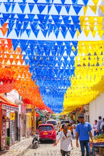 A vibrant street scene with colorful triangular flags hanging overhead in patterns of blue, orange, and yellow. The street is bustling with people walking and a few vehicles parked and moving, including a red car and a motorcycle. Shops line the street, with several signs visible, including one advertising arepas. The atmosphere is lively and bright.