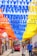 A vibrant street scene with colorful triangular flags hanging overhead in patterns of blue, orange, and yellow. The street is bustling with people walking and a few vehicles parked and moving, including a red car and a motorcycle. Shops line the street, with several signs visible, including one advertising arepas. The atmosphere is lively and bright.