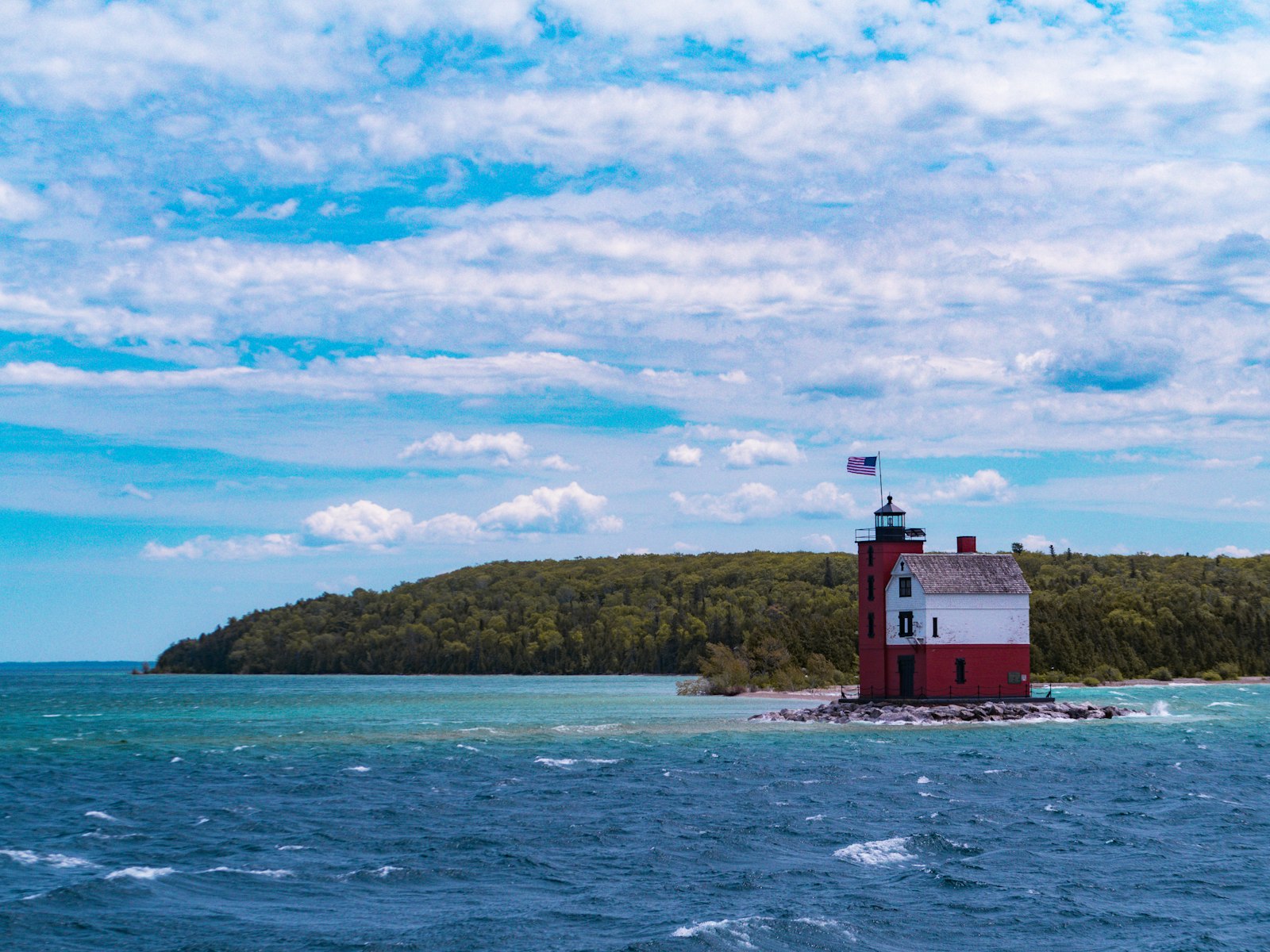 A red-roofed white lighthouse on a rocky point in choppy blue water with a forested island behind it